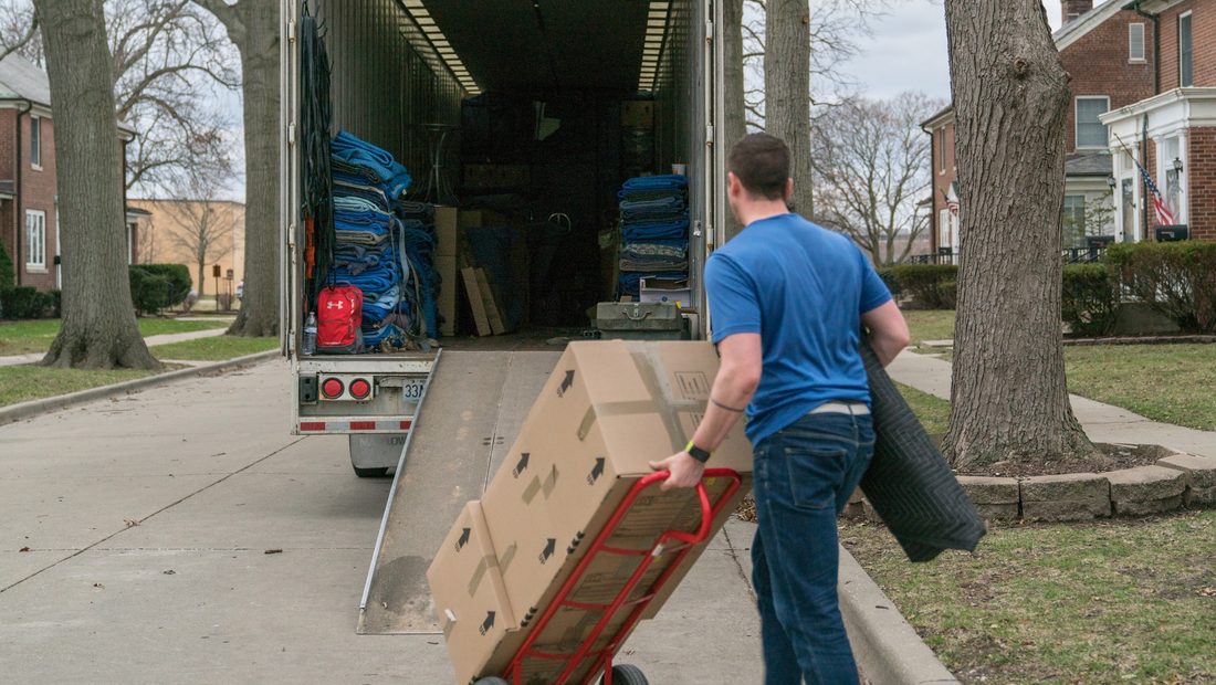 A mover wheels boxes up the ramp of a moving trailer on a street of brick base housing with an American flag on the porch