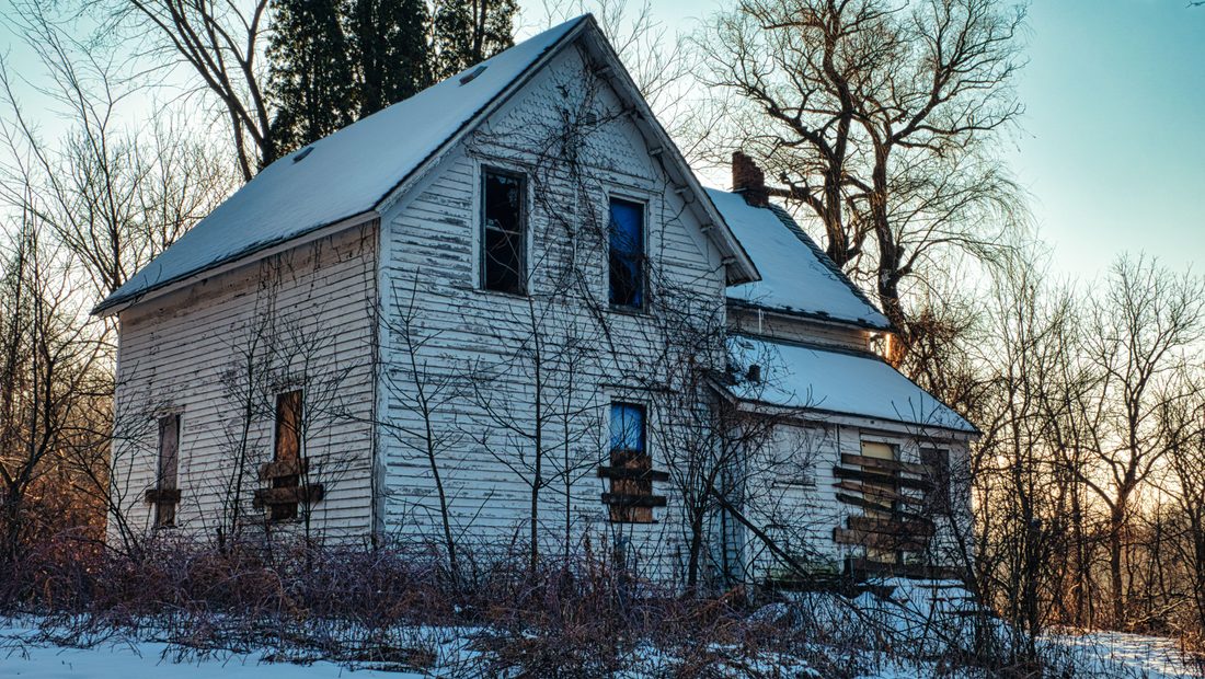 An empty white two-story house in deep snow at dusk with boarded lower windows and dark upper windows